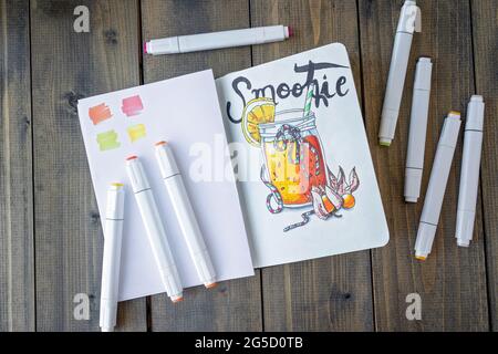 Disposizione piatta con frullato per schizzi nel notebook. Il tutto posto su un taccuino e pennarelli in legno. Hobby. Bicchiere da disegno di frullato. Vista dall'alto. Messa a fuoco morbida Foto Stock