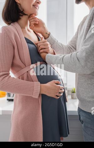vista parziale del giovane uomo che tiene delicatamente il viso e la mano della donna incinta a casa Foto Stock