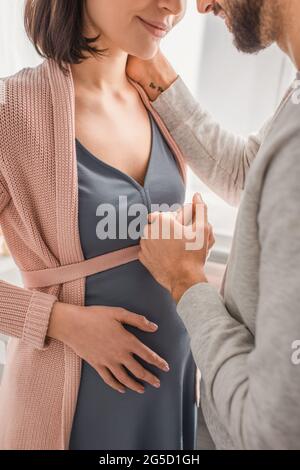 vista parziale del giovane uomo che tiene delicatamente la mano della donna incinta a casa Foto Stock