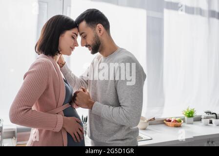 giovane uomo con occhi chiusi che tiene delicatamente la mano della donna incinta in cucina Foto Stock