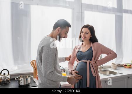 sorridente giovane uomo che tocca delicatamente donna incinta in cucina Foto Stock