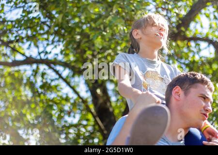 Fratello disinnescato che cavalcava la sorella sulla schiena. Ritratto di ragazza felice sulle spalle dell'uomo, piggyback. La ragazza vola. La famiglia gioca all'aperto. Albero verde Foto Stock