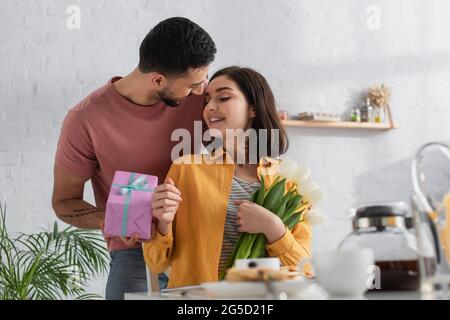 giovane sorridente che presenta una confezione regalo alla fidanzata con bouquet di fiori in cucina Foto Stock