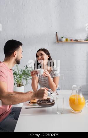 giovane donna sorridente con tazza di caffè guardando il ragazzo con caffettiera in cucina Foto Stock