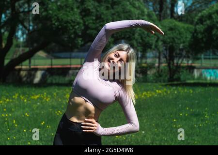 Giovane donna caucasica dai capelli biondi in abbigliamento sportivo sta facendo yoga nel parco. Allenamento all'aperto. Lo yoga pone Foto Stock