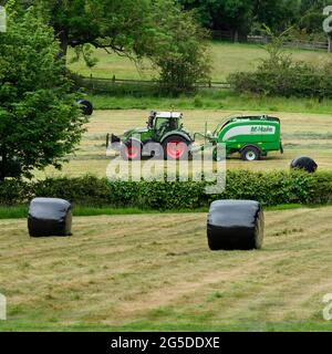 Produzione di fieno o insilato (coltivatore in trattore agricolo al lavoro in campo rurale che traina rotopressa, raccogliendo erba secca e balle rotonde avvolte - Yorkshire Inghilterra, Regno Unito. Foto Stock