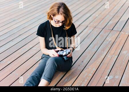 Donna hipster di bellezza con gli occhiali usa la macchina fotografica retrò mentre si siede sul pavimento di legno all'aperto Foto Stock