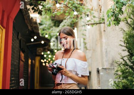 Ritratto di giovane donna turistica appassionata con fotocamera retrò in mano mentre si posa alla vecchia architettura urbana. Scopri nuovi luoghi Foto Stock