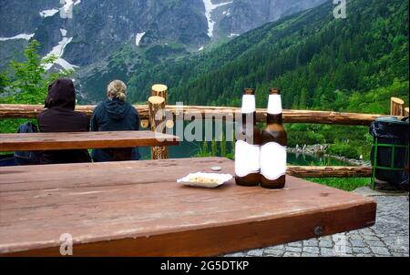 Due bottiglie di bevanda alcolica senza marchio con etichetta bianca accanto agli spuntini sul tavolo di legno contro una coppia di persone seduti su una panchina che si ammira e lo Foto Stock