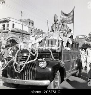 1950s, storico, celebrazione di nozze, Benares, India, Una coppia appena sposata seduta sul tetto di un furgone Chevrolet con cornice in legno con il cofano coperto da ghirlande fiorite decorate o trecce. Foto Stock