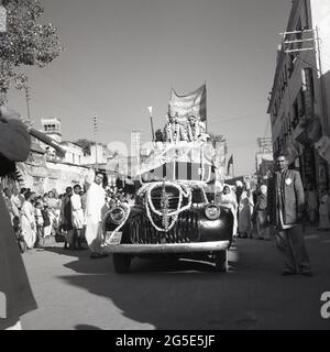 1950s, storico, celebrazione di nozze, Benares, India, Una coppia appena sposata seduta sul tetto di un furgone Chevrolet con cornice in legno con il cofano coperto da ghirlande fiorite decorate o trecce che viaggiano lentamente lungo una strada trafficata. Foto Stock