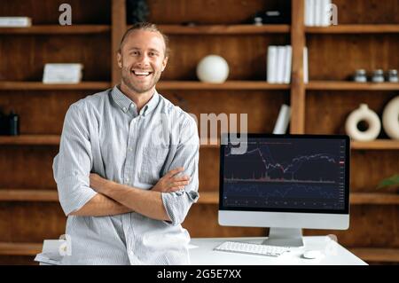 Investimenti, negoziazione in borsa. Positivo successo caucasico uomo trader investitore, si trova vicino scrivania con il computer, guarda la fotocamera, sorridente, trading dati grafico indice sullo schermo del pc Foto Stock