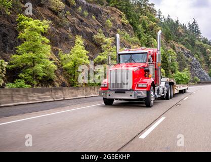 Classico trattore semi-camion industriale con carro grande e potente rosso con cabina corta per il trasporto di un semirimorchio in discesa vuoto che corre su un'alta montagna divisa Foto Stock