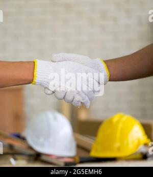 Carpentieri con guanto di protezione che scuotono le mani dopo aver terminato il lavoro. Un casco di costruzione bianco e giallo è stato posto sul tavolo sullo sfondo. Foto Stock