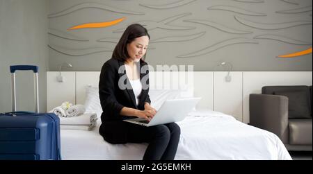 La giovane donna asiatica in abito nero usa il computer portatile per lavorare la mattina a letto. Il suo bagaglio posto accanto al letto. Atmosfera di lavoro fuori du Foto Stock