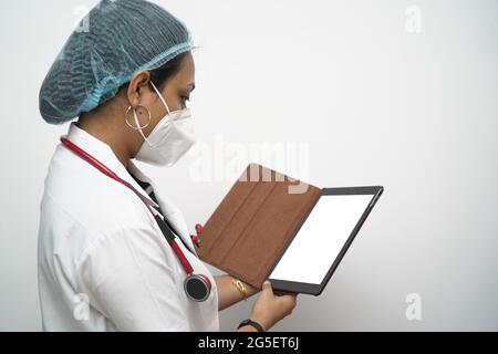 Una dottoressa sudindiana in 30s controllare mockup compressa in camice bianco e stetoscopio rosso su sfondo bianco. Foto Stock