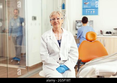 Ortodontista anziano in uniforme medica seduto sulla sedia guardando in macchina fotografica in attesa di uomo pacient per iniziare il trattamento stomatologico dopo l'intervento dentistico. Team di dentisti che lavorano in ufficio Foto Stock