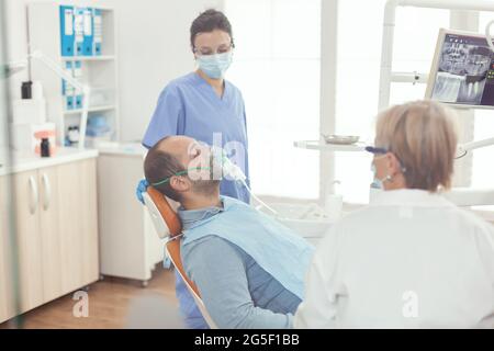 Assistente ospedaliero che mette maschera di oxigen prima di chirurgia dentale seduta su sedia di stomatologia in clinica di odontoiatria . Medico ortodontista senior e infermiere che lavorano in un ufficio moderno indossando maschera e guanti Foto Stock