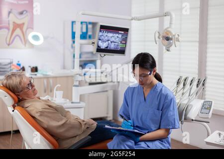 Assistente dentale che completa l'interrogante durante il controllo orale del paziente anziano. Donna anziana che parla con l'infermiera medica in ufficio di stomatologia circa il problema dei denti. Foto Stock