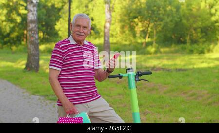 Vecchio nonno caucasico anziano appoggiato su scooter elettrico dopo aver fatto acquisti con borse colorate Foto Stock