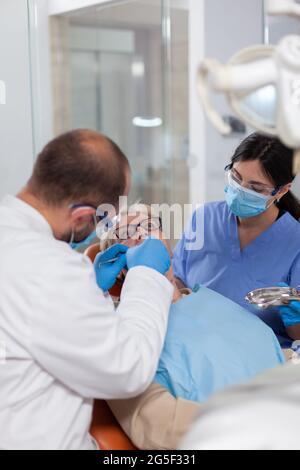 Dentista con assistente installare l'impianto nella clinica moderna della moutina del paziente. Donna anziana durante l'esame medico con stomatolog in studio dentistico con attrezzature arancioni. Foto Stock