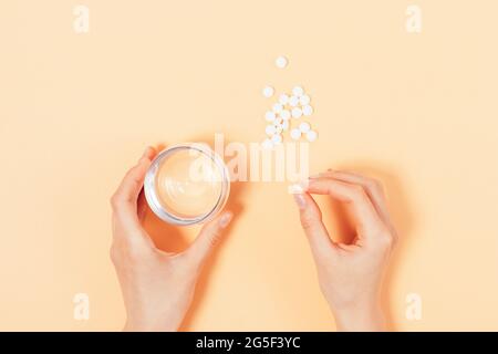 Le mani femminili tengono la pillola e il bicchiere d'acqua accanto al mazzo di compresse su sfondo beige, vista dall'alto. Foto Stock