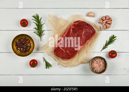 Primo piano su una bistecca di manzo di sidro crudo marmorizzato invecchiato su carta marrone pergamena che avvolge, con spezie, su sfondo bianco tavolo di legno, elevato a. Foto Stock