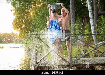 Una coppia anziana si diverte dopo la sauna finlandese sul molo di un cottage in legno in un lago. Uomo maturo che versa acqua fredda su sua moglie. Tipica estate finlandese. Foto Stock