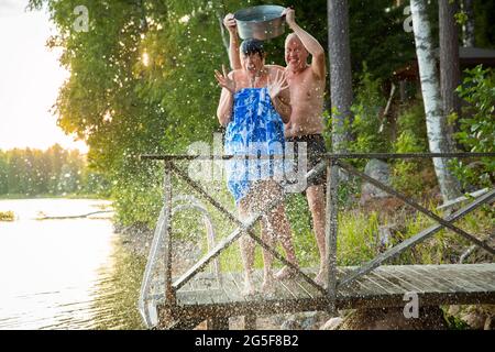 Una coppia anziana si diverte dopo la sauna finlandese sul molo di un cottage in legno in un lago. Uomo maturo che versa acqua fredda su sua moglie. Tipica estate finlandese. Foto Stock