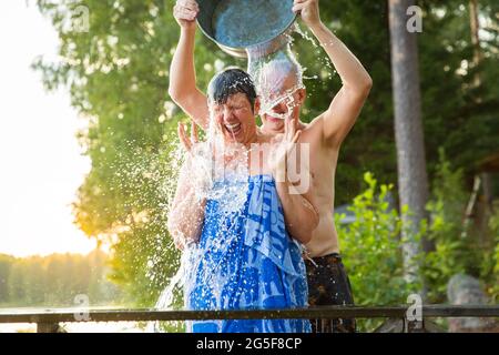 Una coppia anziana si diverte dopo la sauna finlandese sul molo di un cottage in legno in un lago. Uomo maturo che versa acqua fredda su sua moglie. Tipica estate finlandese. Foto Stock