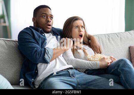 Film horror. Scossa coppia multiculturale guardando film spaventoso e mangiare popcorn a casa, Ritratto di terrorizzati sposi Interrazziale seduto sul divano Foto Stock