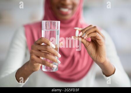 Unriconoscable Donna musulmana nera in Hijab prendendo pillola supplementare e vetro di tenuta dell'acqua, sorridente la signora islamica africana usando le vitamine per sano Foto Stock