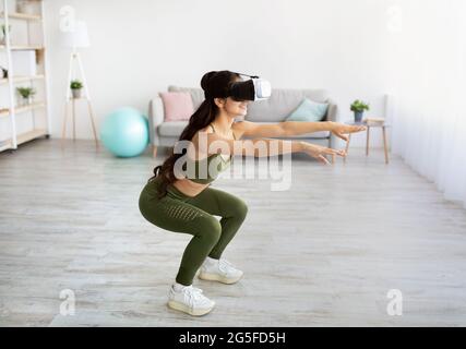 Allenamento domestico con realtà virtuale. Giovane donna indiana con visore VR che fa squat a casa. Atletica millenaria asiatica signora utilizzando tecnologie moderne per Foto Stock