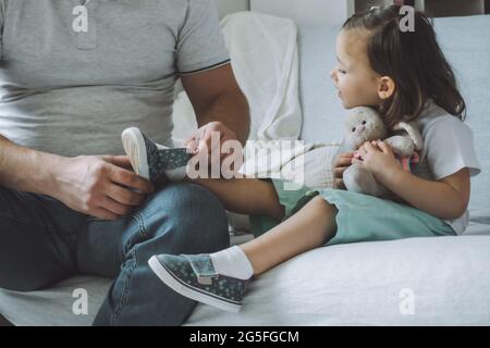 Papà e figlia seduti sul divano. Padre aiuta il bambino a decollo o a indossare scarpe, sorrisi di capretto Foto Stock