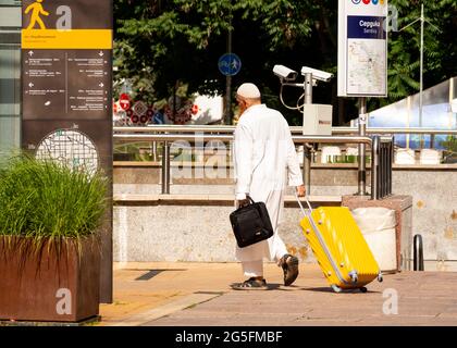 Uomo musulmano di mezza età che indossa un abito bianco e un cappuccio kufi con una cassa di lapton che tira la valigia gialla per le strade di Sofia, Bulgaria Foto Stock