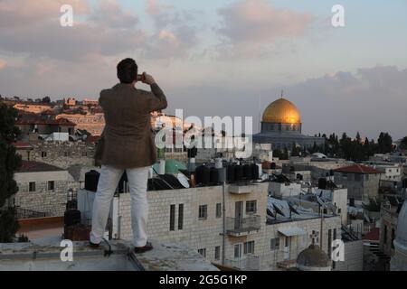 Il turista che si erge sul bordo di un tetto della Città Vecchia fotografa la cupola della roccia, i tetti della Città Vecchia e il Monte Ulivi al tramonto, Gerusalemme, Israele. Foto Stock