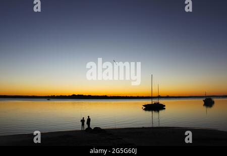 Silhouette di due pescatori a riva e barche a vela in acqua al tramonto Foto Stock