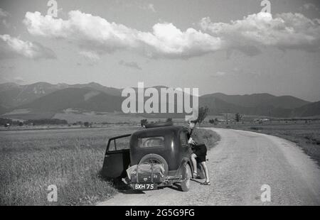 1930s, storico, touring Czechoslavia pre-WW2, giovane uomo in piedi su una strada di campagna, con la sua auto britannica, un Morris 8, che guarda sopra il paesaggio circostante. Dopo il WW1 e la costituzione di uno stato indipendente della Cecoslovacchia, diverse zone di confine del paese, compresa la storica zona occidentale della Boemia, erano ora occupate da una maggioranza di persone di lingua tedesca, nota con il nome storico tedesco come Sudentenland. Il 1938 ha visto la crisi di Sudeten, l'accordo di Monaco e poi il WW2. Foto Stock