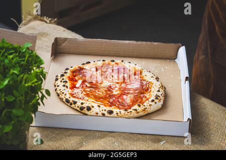 Pizza appena sfornata con salsiccia di salame in scatola di cartone su tela piana. Cibo da asporto, concetto di consegna pizza. Foto Stock