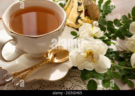 Tazza di tè e rametto di piccole rose bianche su tavola rustica, in colori bianco e oro Foto Stock