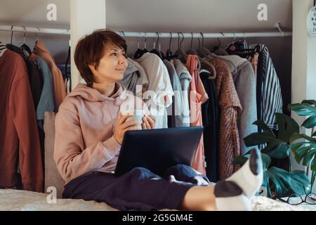 Donna con una tazza di caffè seduto sul letto e utilizzando il computer portatile e godersi il momento. Appartamento con armadio aperto. Casa accogliente. Comunicazione online Foto Stock