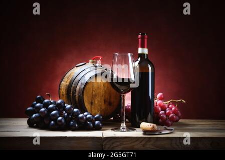 Bicchiere di vino rosso con grappoli d'uva, bottiglia e barile, ancora vivo su tavola di legno. Sfondo rosso, spazio per il testo. Foto Stock