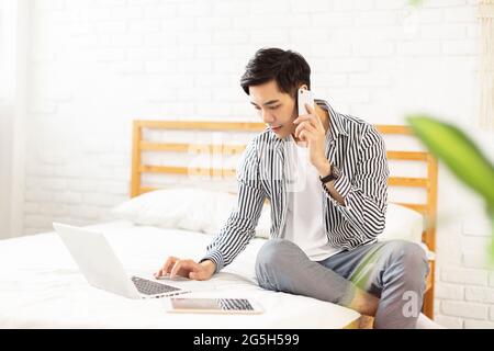 giovane uomo in abiti casual e che lavora a casa mentre si siede sul letto con un computer portatile Foto Stock