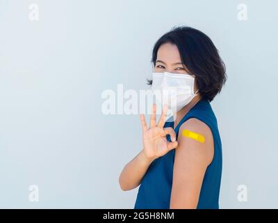 Vaccinazioni, bendaggi sul concetto di braccio delle persone vaccinate. Felice donna asiatica d'affari che indossa giubbotto blu blazer, maschera viso mostrando bendaggio giallo e ok Foto Stock