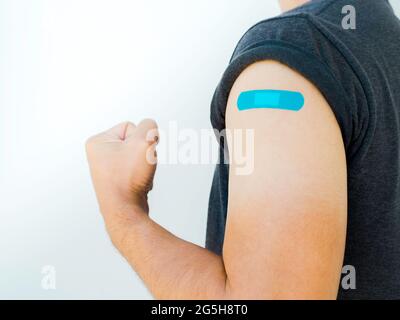 Vaccinazioni, bendaggio su persone vaccinate concetto. Bendaggio blu sulla spalla dell'uomo che indossa una camicia grigia e mostra un gesto forte con pugno e braccio A. Foto Stock
