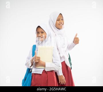 sorridendo due ragazze velate che indossano uniformi scolastiche elementari con i pollici in su trasportano gli zaini e un libro Foto Stock