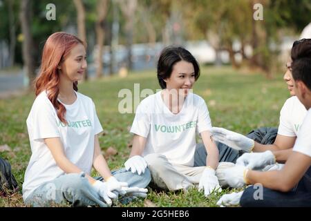 Gruppo di giovani volontari sorridenti che si siedono sull'erba e parlano dopo aver raccolto i rifiuti nel parco cittadino Foto Stock