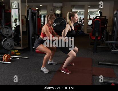 Allenatore personale femminile che aiuta la giovane donna che fa squats barbelll in palestra Foto Stock