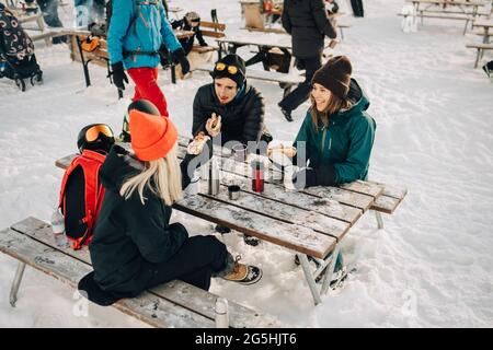Amici di sesso maschile e femminile che parlano mentre mangiano il cane caldo nella stazione sciistica durante l'inverno Foto Stock