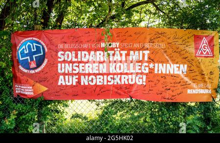 Rendsburg, Germania. 28 Giugno 2021. I sindacalisti di IG Metall hanno appeso un poster con l'iscrizione "solidarietà con i nostri colleghi di Nobiskrug" con innumerevoli firme di fronte all'ingresso del cantiere Nobiskrug e posto 155 sedie davanti all'ingresso del cantiere. Con lo slogan 'non ci sono più sedie! Lavoro e futuro per Nobiskrug!', IG Metall attira l'attenzione sul licenziamento di 155 dipendenti. Ora si tratta di preservare i restanti 300 posti di lavoro, ha detto l'amministratore delegato di IG Metall Rendsburg. Credit: Axel Heimken/dpa/Alamy Live News Foto Stock